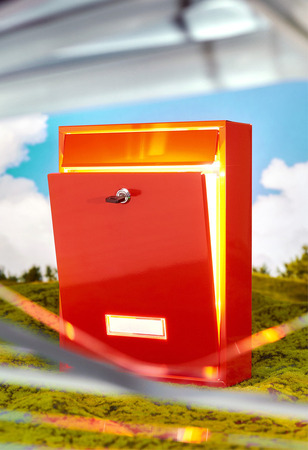 Ein roter Briefkasten steht vor einem Hintergrund aus blauem Himmel und grüner Landschaft. Der Briefkasten hat einen Schlitz für Post und ein Schloss. Die Umgebung wirkt wie eine Miniaturlandschaft.