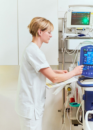Eine Krankenschwester in einem Krankenhauszimmer steht vor einem medizinischen Gerätewagen. Sie trägt einen weißen Kittel und liest in einem Ordner. Im Hintergrund ist ein Monitor mit Vitalwerten zu sehen. Die Atmosphäre wirkt konzentriert und professionell.