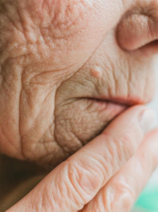 Nahaufnahme eines älteren Menschen, dessen Hand das eigene Gesicht berührt. Die Haut weist viele Falten auf, was auf ein hohes Alter hindeutet. Der Blick ist geschlossen, was Trauer oder Nachdenklichkeit suggeriert.