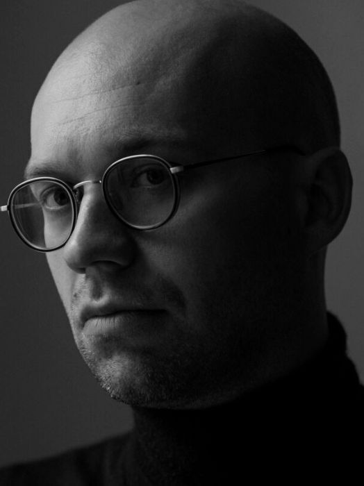 Schwarz-Weiß Porträt eines Mannes mit Glatze und Brille. Er blickt direkt in die Kamera, sein Gesichtsausdruck wirkt ernst und nachdenklich. Er trägt einen dunklen Rollkragenpullover, der Hintergrund ist dunkel gehalten.