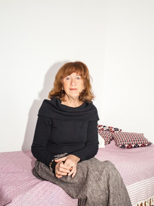 Eine ältere Frau mit roten, lockigen Haaren sitzt auf einem Bett. Sie trägt einen dunklen Pullover mit weitem Ausschnitt und eine graue Hose. Ihre Hände sind in ihrem Schoß gefaltet. Sie blickt direkt in die Kamera und wirkt freundlich und nachdenklich. Im Hintergrund ist ein weißer Wandabschnitt und ein Teil eines Bettes mit gemusterten Kissen zu sehen.