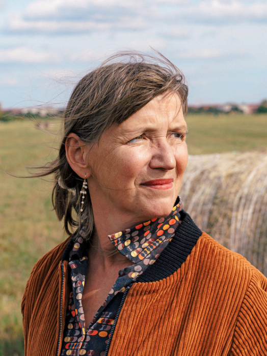 Porträt einer Frau mittleren Alters mit freundlichem Gesichtsausdruck. Sie hat graues, leicht zerzaustes Haar und trägt eine orangefarbene Cordjacke über einem gemusterten Oberteil. Im Hintergrund ist ein Feld zu sehen.