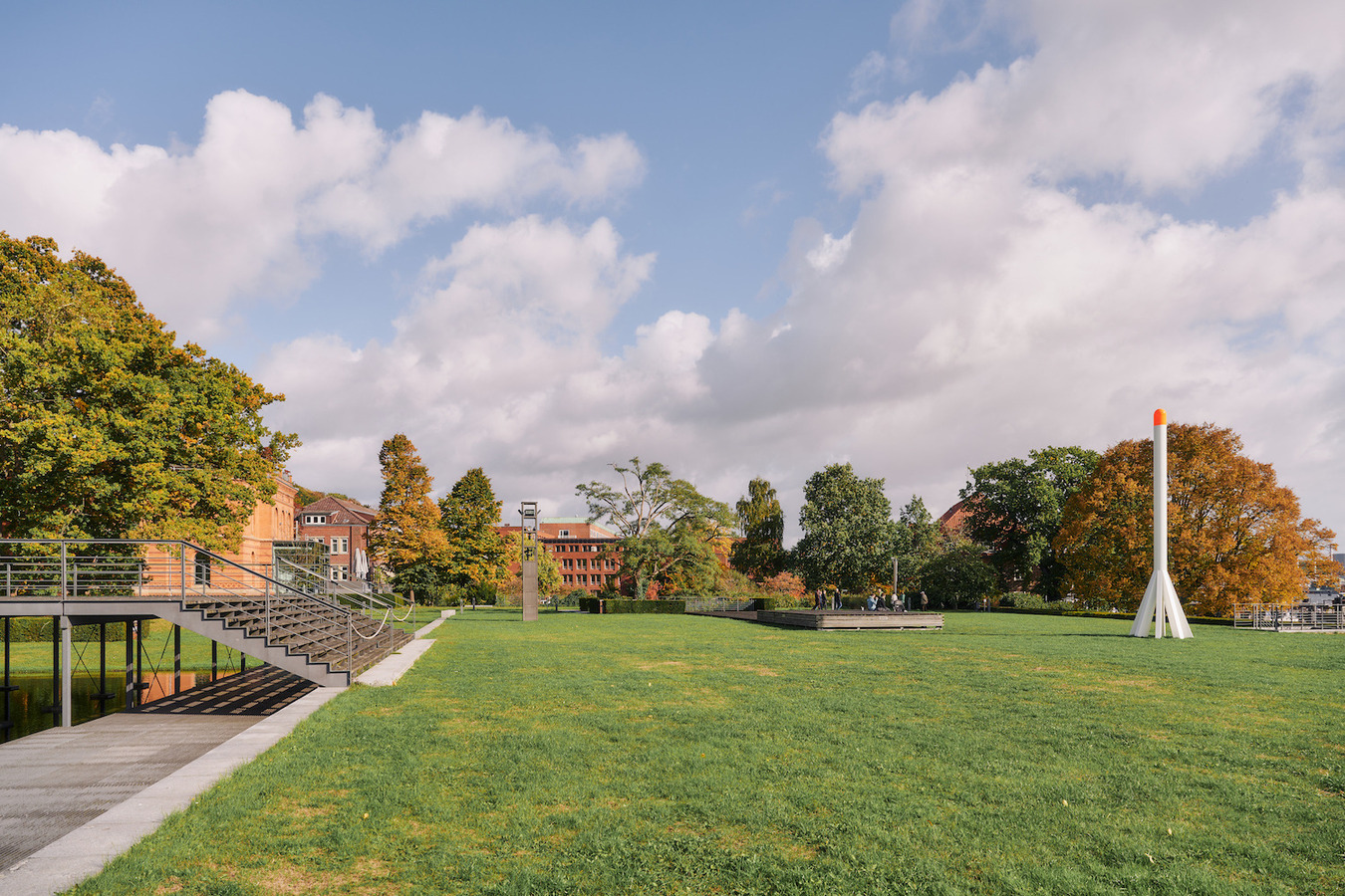 Ein weitläufiger Park mit gepflegtem Rasen. Im Hintergrund ein Gebäude mit roten Ziegeldächern und Bäumen in Herbstfarben. Links führt eine Treppe zu einem Wasserbecken. Rechts steht eine moderne Skulptur in Orange und Grau.