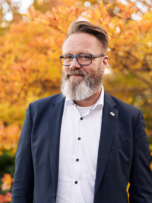 Ein Mann mit grau meliertem Vollbart und Brille lächelt freundlich in die Kamera. Er trägt ein weißes Hemd und einen dunkelblauen Blazer. Im Hintergrund verschwimmt ein Herbstlaub in warmen Orange- und Gelbtönen.