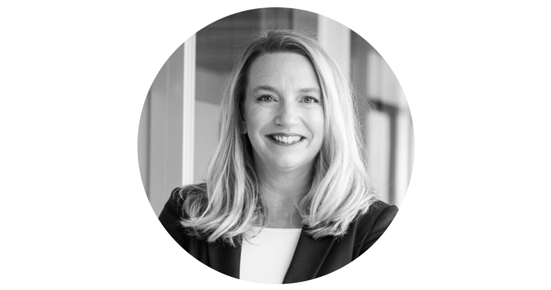 Schwarz-Weiß Porträt einer Frau mittleren Alters. Sie hat blonde Haare und ein freundliches Lächeln. Sie trägt einen Blazer und ein helles Oberteil. Der Hintergrund ist unscharf und deutet auf einen Innenraum hin.
