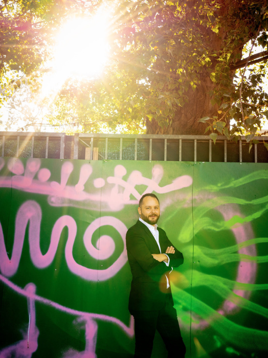 Ein Mann mit Bart steht selbstbewusst vor einer grauen Wand mit grüner Graffiti-Schrift. Er trägt einen dunklen Anzug und einen weißen Schal. Das Licht scheint hell von oben und erzeugt einen starken Kontrast.