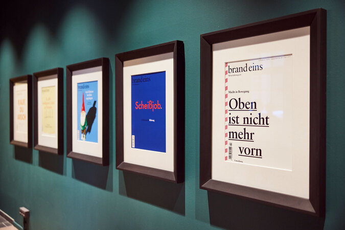 Fünf gerahmte Zeitschriften-Cover hängen an einer dunkelgrünen Wand. Die Cover sind in schwarzen Rahmen gefasst und scheinen Teil einer Ausstellung zu sein. Das Cover am rechten Bildrand ist näher und zeigt den Titel "brand eins" und den Untertitel "Oben ist nicht mehr vorn".