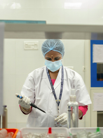 Eine Frau in einem weißen Laborkittel, Mundschutz und Haarnetz arbeitet konzentriert in einem Labor. Sie hält eine Pipette in der Hand und scheint eine Flüssigkeit in ein Reagenzglas zu füllen. Im Hintergrund sind Laborgeräte und Regale zu sehen.