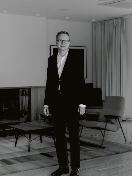 Schwarz-Weiß-Porträt eines Mannes in einem modernen Interieur. Der Mann trägt einen dunklen Anzug und ein helles Hemd, eine Brille und wirkt selbstbewusst und nachdenklich. Er steht in einem Raum mit minimalistischer Einrichtung, darunter ein Sessel und ein Couchtisch. Das Bild vermittelt eine ruhige und elegante Atmosphäre.