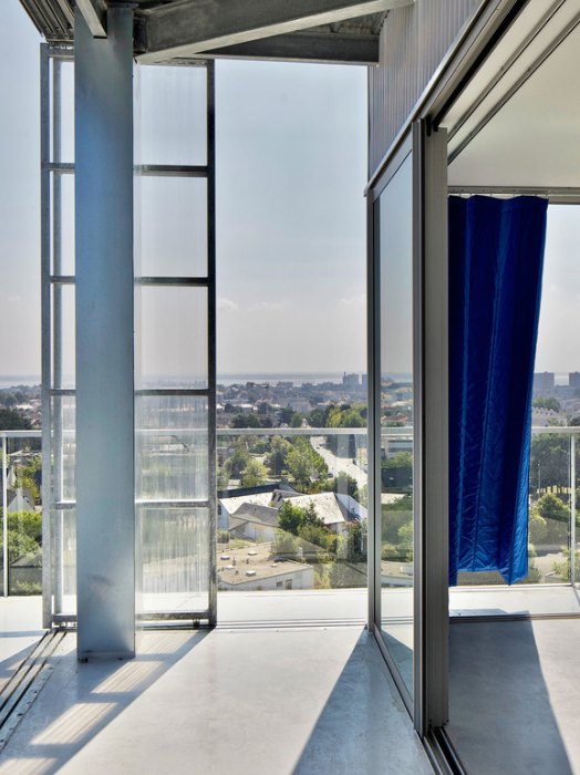 Blick aus einem modernen Gebäude auf eine städtische Landschaft. Große, offene Fensterfronten geben den Blick frei auf eine weitläufige Stadt mit vielen Gebäuden und Bäumen. Ein blauer Stoff hängt links im Bild herunter und sorgt für einen Farbkontrast. Das Ambiente wirkt hell und weitläufig.