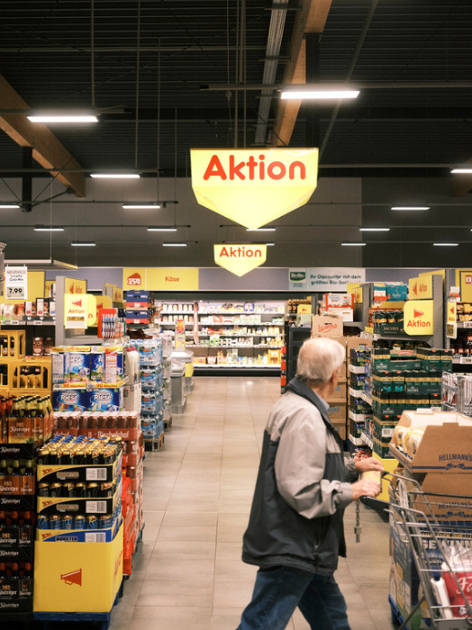 Ein älterer Mann schiebt einen Einkaufswagen durch einen hell erleuchteten Aktions-Markt. Regale voller Waren säumen den Gang, im Hintergrund eine Kühltheke. Das Bild vermittelt ein Gefühl von alltäglichem Einkaufen.
