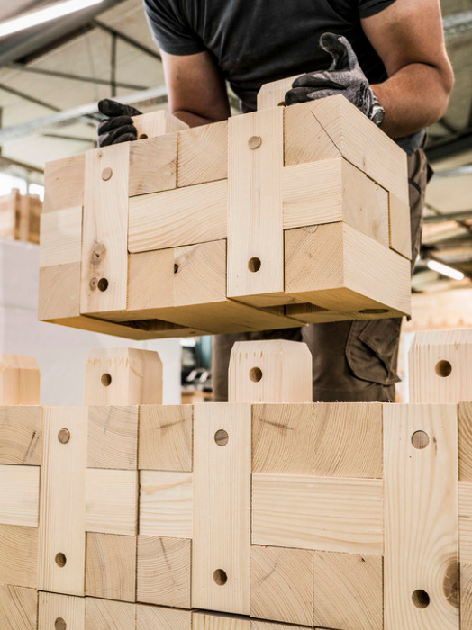 Ein Mann in dunkler Arbeitskleidung und Handschuhen stapelt große Holzbausteine übereinander. Die Bausteine sind helles Holz und haben Aussparungen für eine einfache Verbindung. Im Hintergrund sind weitere gestapelte Holzbausteine zu sehen, vermutlich in einer Werkstatt oder Lagerhalle.