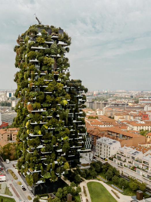 Ein hoher Wohnkomplex, der vollständig mit Bäumen und Pflanzen bewachsen ist. Der Turm steht in einer städtischen Umgebung mit weiteren Gebäuden und Straßen im Hintergrund. Das Ambiente wirkt modern und grün.