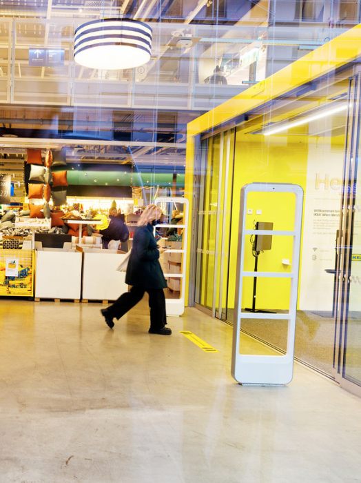 Ein Mann in einem dunklen Mantel geht durch einen hellen IKEA-Markt. Im Hintergrund sind Regale mit Waren und ein gelber Bereich mit einem Informationsterminal zu sehen. Das Bild wirkt durch Bewegungsunschärfe dynamisch.