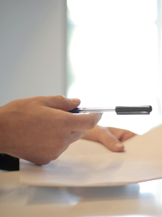 Eine Person hält einen schwarzen Stift über ein weißes Blatt Papier, bereit zu schreiben. Im Hintergrund ist ein Fenster zu sehen, das helles Licht hereinlässt. Die Situation deutet auf eine konzentrierte Tätigkeit wie Notizen machen oder Unterschreiben hin.
