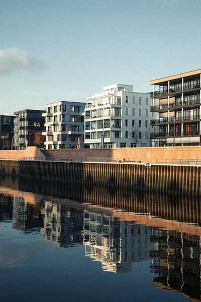 Ein ruhiges Stadtbild am Wasser. Moderne Wohngebäude mit Balkonen reihen sich an einer Uferpromenade aneinander. Die Fassaden spiegeln sich im dunklen Wasser wider. Der Himmel ist hellblau und leicht bewölkt, was eine friedliche Atmosphäre schafft.