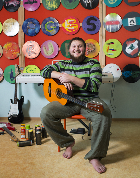 Ein lächelnder Mann mit Bart sitzt auf einem orangefarbenen Stuhl und hält eine Gitarre im Arm. Im Hintergrund eine rote Wand, die mit zahlreichen bemalten Schallplatten bedeckt ist. Links stehen ein Keyboard und Spraydosen auf dem Boden.