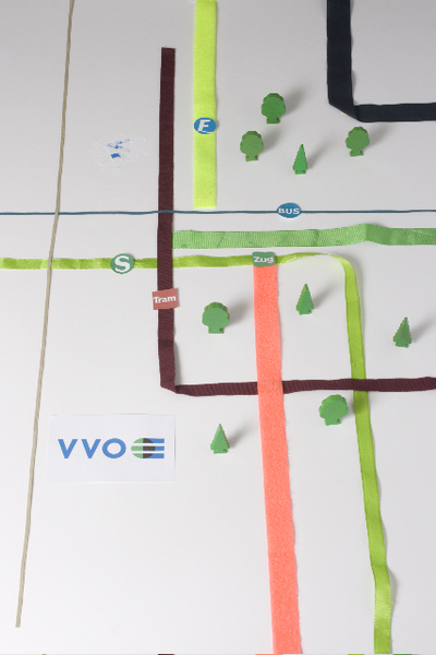 Es handelt sich um eine vereinfachte grafische Darstellung eines öffentlichen Verkehrsnetzes. Mehrere farbige Linien – rot, grün, blau, violett und braun – verlaufen kreuz und quer über einen hellgrauen Hintergrund. An einigen Kreuzungspunkten sind kleine grüne Bäume abgebildet. Die Linien sind mit den Verkehrsmitteln "Tram", "Zug" und "Bus" beschriftet. Links unten befindet sich das Logo "VVO".
