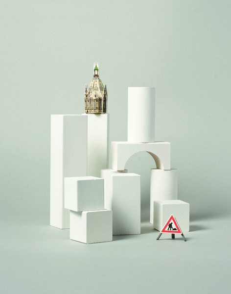 Eine Miniatur-Kathedrale steht auf einem weißen Block, der Teil einer abstrakten Konstruktion aus verschiedenen weißen Formen ist. Vor der Konstruktion befindet sich ein rotes Warndreieck mit einem Fußgänger-Symbol. Der Hintergrund ist ein helles Pastellgrün.