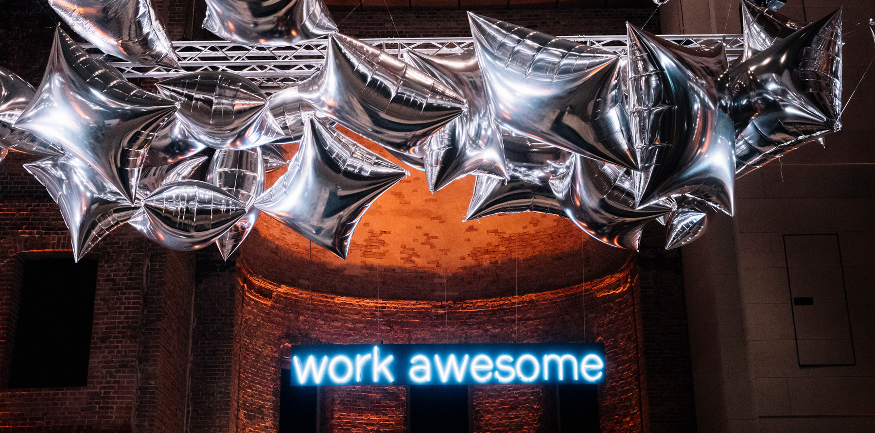 Das Bild zeigt einen dunklen Raum mit einer auffälligen Dekoration aus silbernen Sternballons, die von der Decke hängen. Im Hintergrund ist eine Backsteinwand zu sehen. Ein leuchtendes blaues Neonlicht-Schild mit der Aufschrift "work awesome" befindet sich zentral im Bild und zieht die Aufmerksamkeit auf sich. Die Atmosphäre wirkt festlich und motivierend.