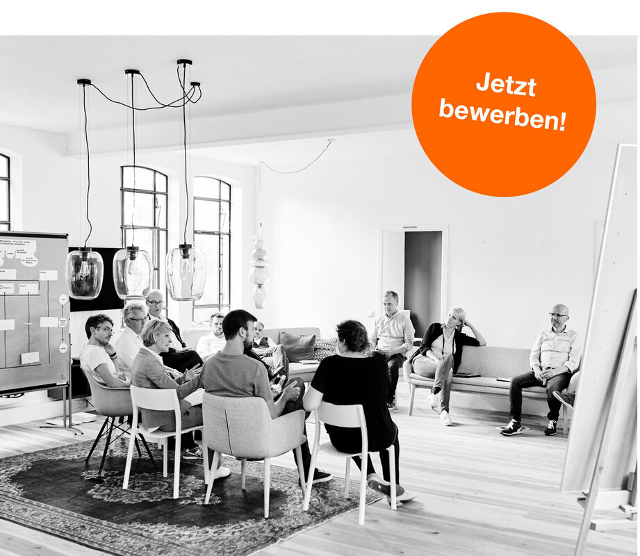 Schwarz-Weiß Aufnahme einer Gruppe von etwa 15 Personen, die um einen großen runden Tisch in einem hellen Raum sitzen. Die Personen scheinen in einer Diskussion oder einem Meeting zu sein. Einige schauen auf Notizen oder den Tisch, andere sind in Gespräche vertieft. Im Hintergrund ist eine Pinnwand mit Papieren zu sehen. Ein orangefarbener Kreis mit der Aufschrift "Jetzt bewerben!" befindet sich rechts im Bild.