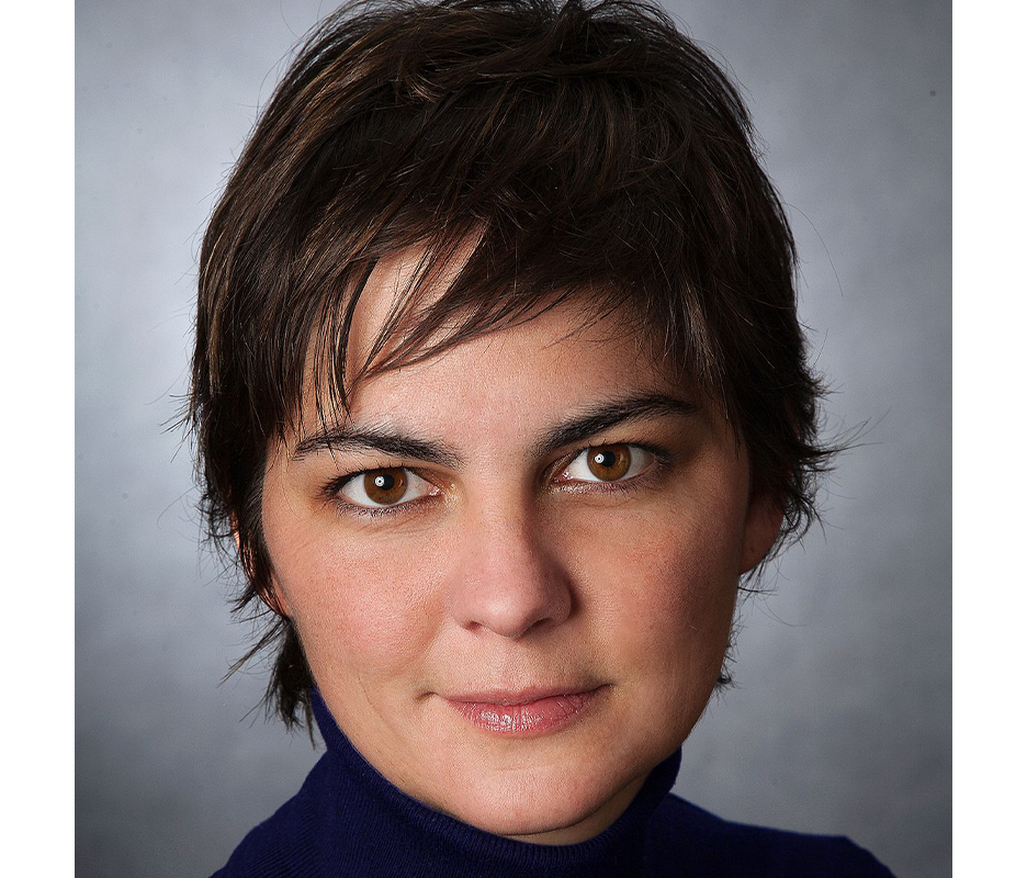 Porträt einer Frau mit kurzen, dunklen Haaren und braunen Augen. Sie trägt einen dunkelblauen Rollkragenpullover und blickt direkt in die Kamera. Ihr Gesichtsausdruck wirkt ruhig und selbstbewusst.