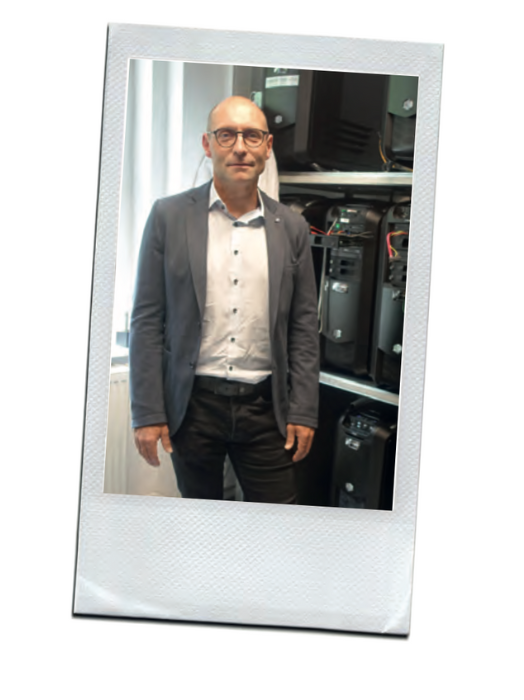 Ein Mann mittleren Alters mit Glatze und Brille steht lächelnd vor einer technischen Einrichtung. Er trägt einen grauen Blazer und ein helles Hemd. Im Hintergrund sind Server oder Computer zu sehen, was auf eine Büroumgebung hindeutet.