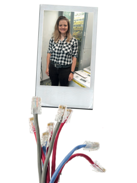 Eine junge Frau mit blonden Haaren steht lächelnd vor einem Serverraum. Sie trägt ein kariertes Hemd und eine dunkle Hose. Unter ihr befinden sich mehrere Netzwerkkabel in verschiedenen Farben.