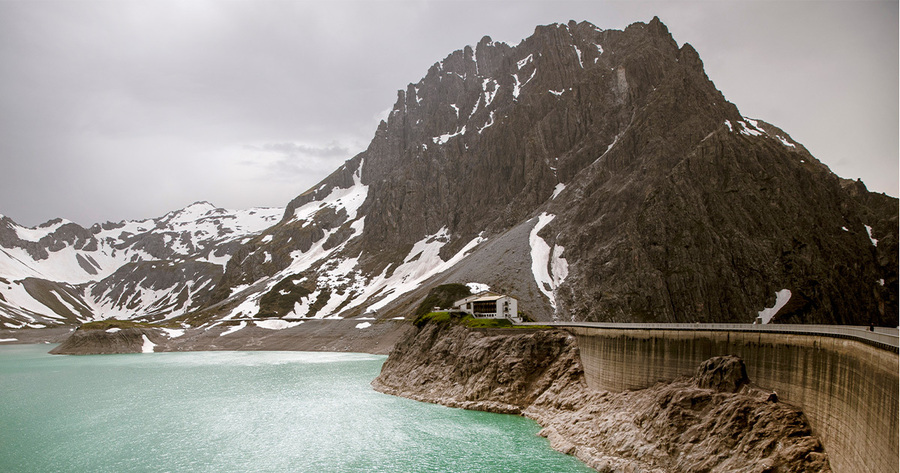 Ein See unterhalb eines hohen, zerklüfteten Berges mit Schneeflecken. Am Ufer des Sees befindet sich eine Staumauer mit einer Straße darauf. Der Himmel ist bewölkt, was eine ruhige und etwas düstere Atmosphäre erzeugt.