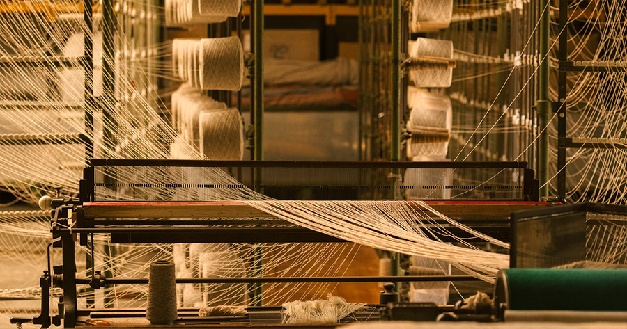 Das Bild zeigt eine Webmaschine in einer Fabrikhalle. Viele Fäden erstrecken sich in einem komplexen Muster über die Maschine. Das Licht ist warm und betont die Textur der Fäden und die industrielle Umgebung.
