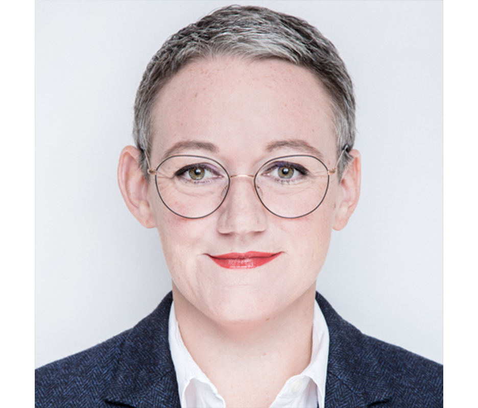 Porträt einer Frau mit kurzem, grauem Haar und runden Brillengläsern. Sie trägt einen dunklen Blazer und ein helles Hemd. Ihr Blick ist freundlich und sie wirkt selbstbewusst.