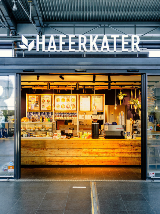 Das Bild zeigt die Frontansicht eines Cafés namens "Haferkater". Durch die großen Glasfronten ist der Innenraum gut sichtbar. Dort befindet sich eine Theke mit einer Auswahl an Gebäck und Snacks. Im Hintergrund sind Menüs an der Wand angebracht. Das Ambiente wirkt einladend und modern.