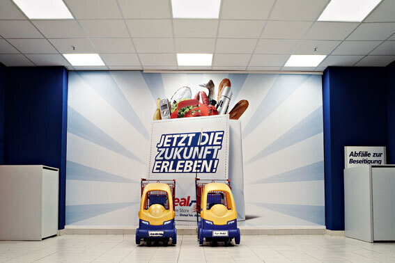 Das Bild zeigt einen hellen Raum, vermutlich ein Geschäft oder eine Wartezone. An der blauen Wand befindet sich ein großes Werbeplakat mit der Aufschrift "Jetzt die Zukunft erleben!". Das Plakat zeigt eine Vielzahl von Lebensmitteln und Konsumgütern, die aus einer Einkaufstasche quellen. Vor dem Plakat stehen zwei gelbe Kinderautos nebeneinander. Rechts im Bild befindet sich ein grauer Schrank mit der Aufschrift "Abfälle zur Beseitigung".