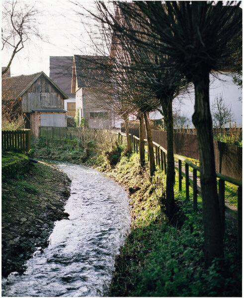 Ein kleiner Bach fließt durch eine ländliche Gegend. Am Ufer stehen Bäume mit nackten Ästen und ein Holzzaun. Im Hintergrund sind Häuser mit Ziegeldächern zu sehen. Das Licht scheint warm und deutet auf einen sonnigen Tag hin.