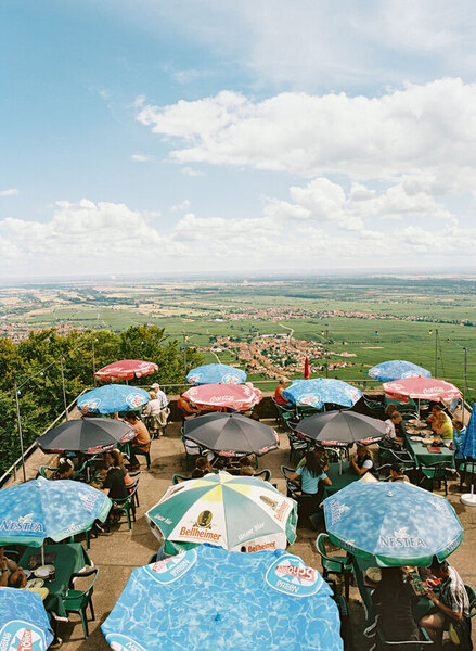 Das Bild zeigt eine Terrasse mit vielen Sonnenschirmen und Sitzgruppen. Im Hintergrund erstreckt sich eine weite Landschaft mit Feldern und einer Stadt. Menschen sitzen an den Tischen und scheinen die Aussicht zu genießen. Es herrscht eine entspannte Atmosphäre.