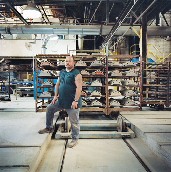 Ein Mann steht selbstbewusst in einer großen Industriehalle. Er hat kurze Haare, einen Bart und trägt ein grünes Tanktop, Jeans und Arbeitsschuhe. Im Hintergrund befinden sich Metallregale mit vielen gleichförmigen Metallteilen auf Rollen. Die Halle wirkt hell und funktional.