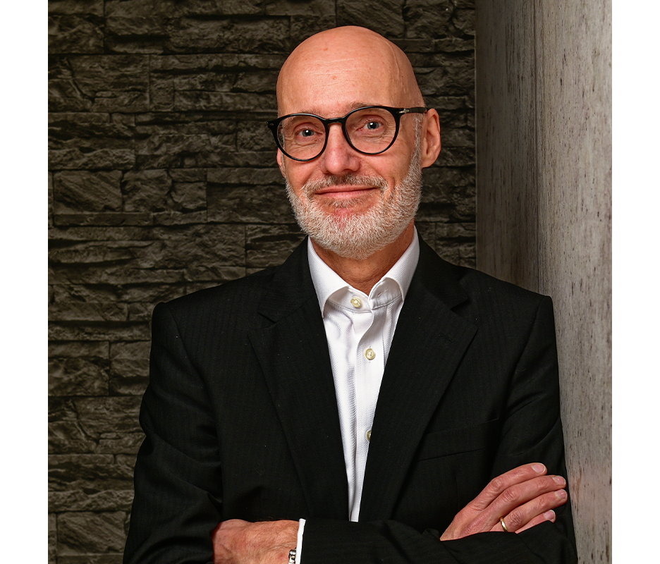 Ein Mann mit Glatze und Vollbart trägt eine Brille und einen dunklen Anzug. Er lächelt freundlich in die Kamera und wirkt sympathisch. Im Hintergrund ist eine graue Steinwand zu sehen.