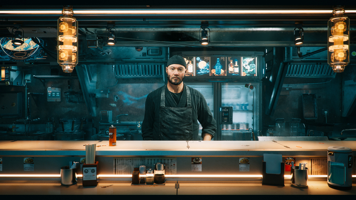 Ein Mann mit dunklem Haar und Bart steht hinter einer Theke in einem dunklen, futuristisch wirkenden Restaurant. Er trägt eine schwarze Schürze und scheint freundlich in die Kamera zu schauen. Im Hintergrund sind Regale und Menüs zu sehen, die von warmem Licht beleuchtet werden. Das Ambiente wirkt modern und einladend.