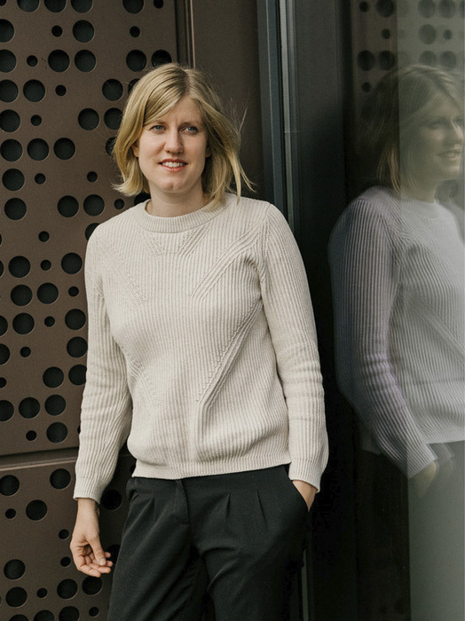Eine junge Frau mit blonden, kurzen Haaren lächelt freundlich in die Kamera. Sie trägt einen beigen Pullover und eine dunkle Hose und steht vor einer Fassade mit einem Muster aus runden Löchern. Ihr Spiegelbild ist im Glas zu sehen.
