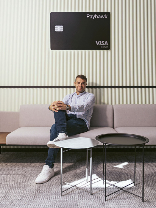 Ein Mann sitzt entspannt auf einem hellrosa Sofa. Er trägt ein hellblaues Hemd und Jeans. Vor ihm steht ein kleiner, runder Tisch aus Metall. Über dem Sofa schwebt eine schwarze Kreditkarte mit dem Aufdruck "Payhawk VISA Platinum". Der Hintergrund ist eine geriffelte, helle Wand. Das Bild vermittelt einen Eindruck von Modernität und finanzieller Sicherheit.
