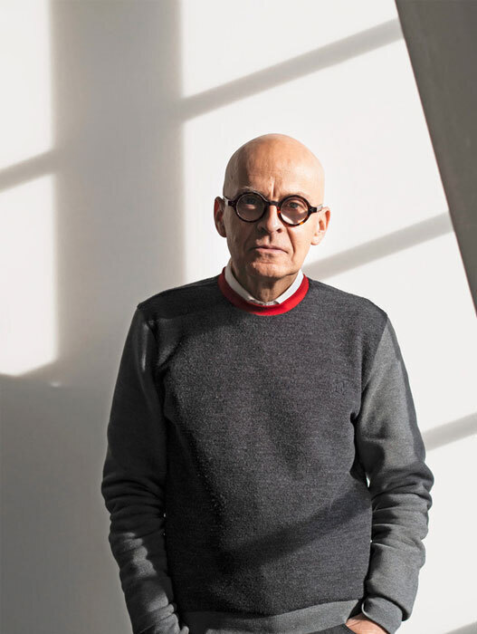 Ein älterer Mann mit Glatze und runder Brille blickt ernst in die Kamera. Er trägt einen grauen Pullover mit rotem Kragen. Das Licht fällt schräg von der Seite auf ihn und erzeugt starke Schatten.