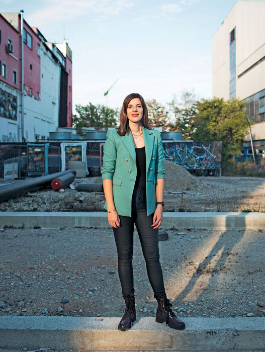 Eine junge Frau steht selbstbewusst in einem urbanen Umfeld. Sie trägt eine türkisfarbene Jacke, eine schwarze Hose und Stiefel. Im Hintergrund sind Gebäude und eine Baustelle zu sehen. Die Frau wirkt entschlossen und modern.