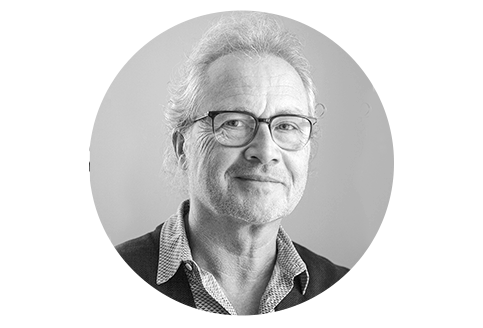 Schwarz-Weiß Porträt eines älteren Mannes mit lockigem, grau meliertem Haar und Brille. Er trägt ein kariertes Hemd und einen dunklen Pullover. Sein Blick ist freundlich und wirkt einladend.