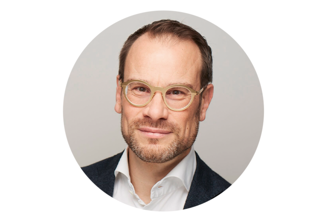 Porträt eines Mannes mit Brille. Er hat kurze braune Haare und einen Vollbart. Er trägt einen dunklen Anzug und wirkt freundlich und zugewandt.