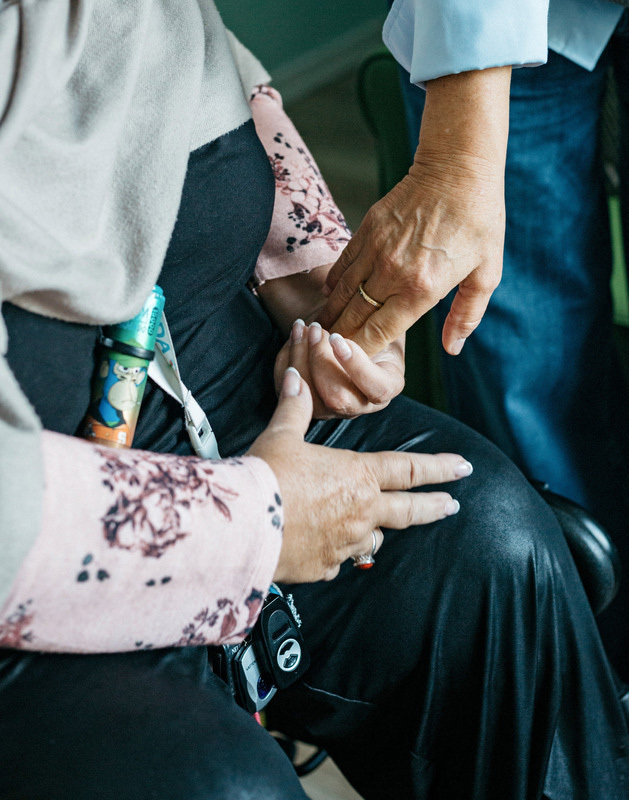 Zwei ältere Hände halten sich fest. Die Person links trägt einen rosafarbenen Pullover mit floralem Muster und eine Smartwatch. Die Person rechts trägt ein hellblaues Hemd. Die Aufnahme zeigt die Hände im Nahbereich, was eine Atmosphäre von Nähe und Unterstützung vermittelt.