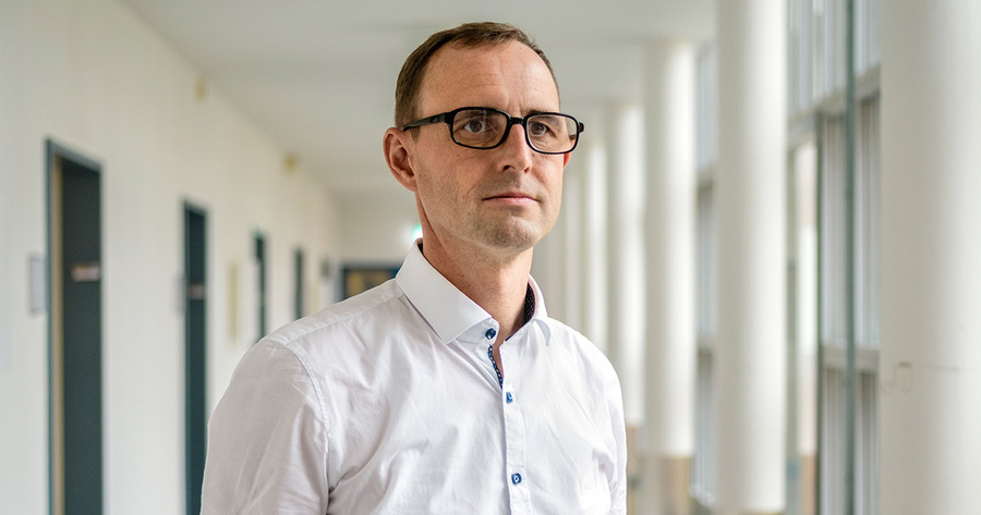 Ein Mann mit kurzgeschnittenem, dunklem Haar und Brille blickt selbstbewusst in die Kamera. Er trägt ein hellblaues Hemd mit offenem Kragen. Im Hintergrund befindet sich ein heller Flur mit Türen und Säulen, der eine Büroatmosphäre suggeriert.