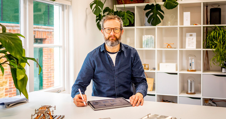Ein Mann mittleren Alters mit Bart sitzt an einem Schreibtisch und zeichnet auf einem Grafiktablett. Er trägt ein blaues Hemd und eine Brille. Er wirkt konzentriert und nachdenklich. Im Hintergrund sind Regale mit Pflanzen und Büchern zu sehen, die eine ruhige Arbeitsatmosphäre schaffen.