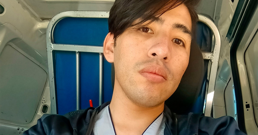 Ein Mann mit dunklen Haaren und braunen Augen blickt direkt in die Kamera. Er hat einen leichten Bartschatten und wirkt nachdenklich. Im Hintergrund ist der Innenraum eines Fahrzeugs zu sehen, vermutlich ein Transporter oder Van, mit blauen Metallstreben.