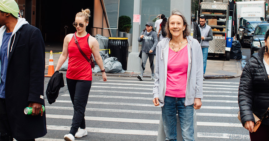 Das Bild zeigt eine belebte Straßenszene. Mehrere Personen überqueren eine Straße. Im Vordergrund eine Frau mittleren Alters mit lockigem Haar, die lächelt und freundlich wirkt. Sie trägt Jeans und einen rosa Pullover. Neben ihr geht eine jüngere Frau mit Sonnenbrille und rotem Top. Im Hintergrund sind weitere Fußgänger und ein Imbisswagen zu sehen.
