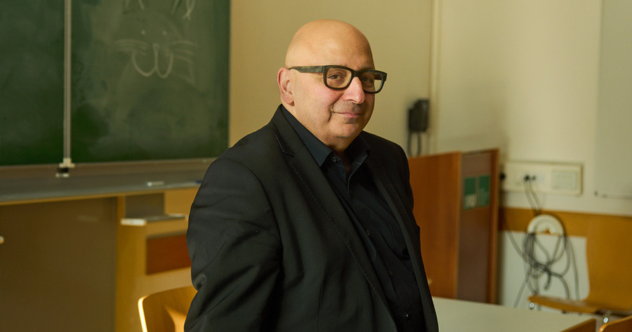 Ein Mann mit Glatze und Brille lächelt freundlich in die Kamera. Er trägt einen dunklen Anzug und befindet sich in einem Klassenzimmer, im Hintergrund sind Tische und eine Tafel zu sehen.